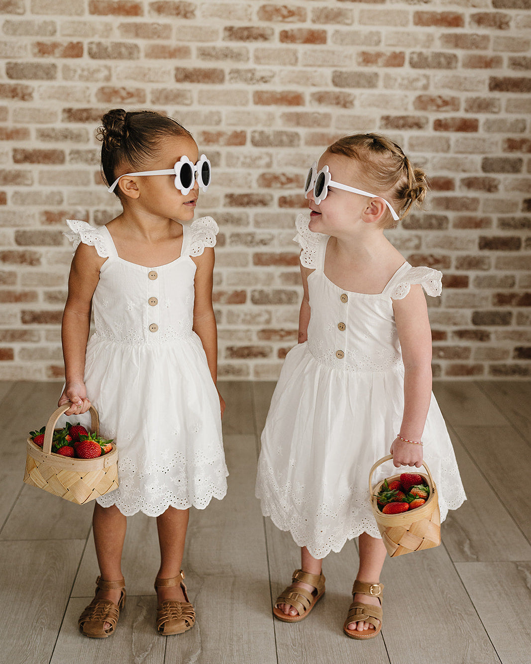 Two young girls in white dresses standing against a brick wall.
