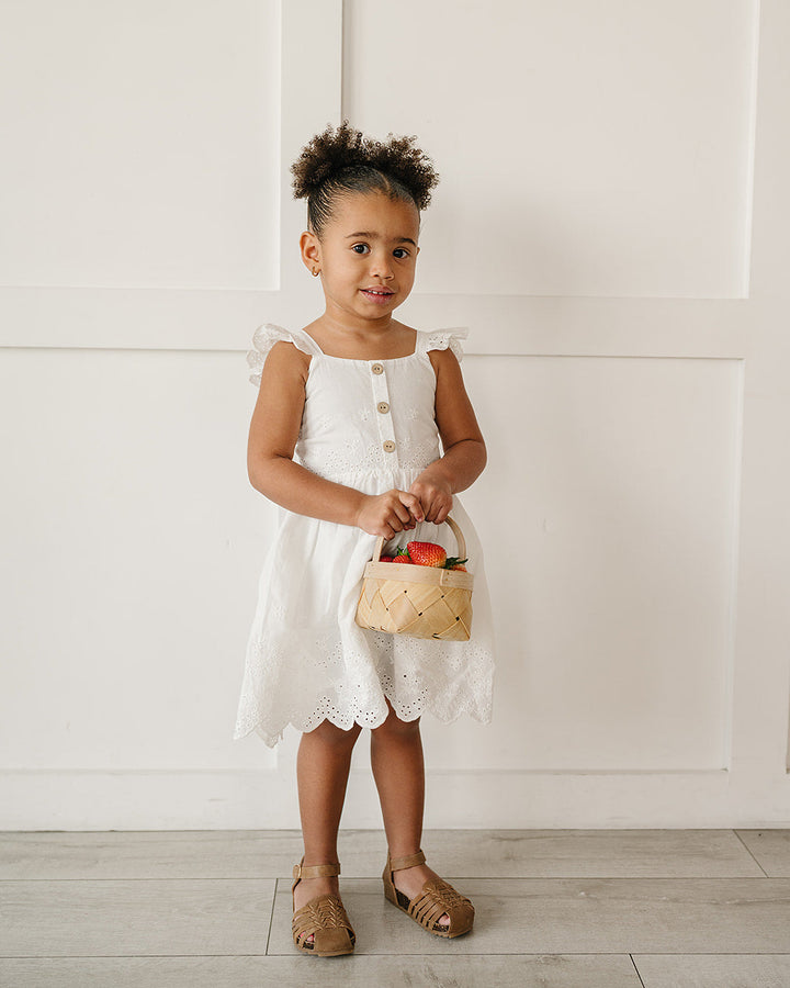 Young girl in a white dress holding a basket with flowers against a white wall.
