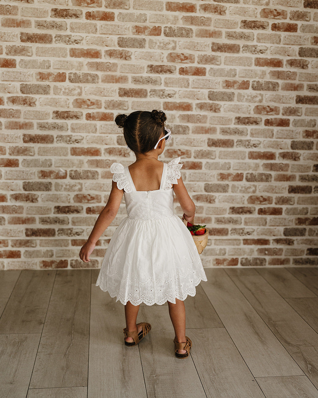 Young girl in a white dress standing against a brick wall.