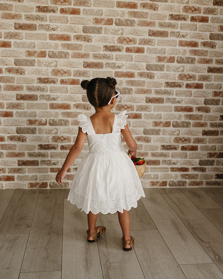 Young girl in a white dress standing against a brick wall.