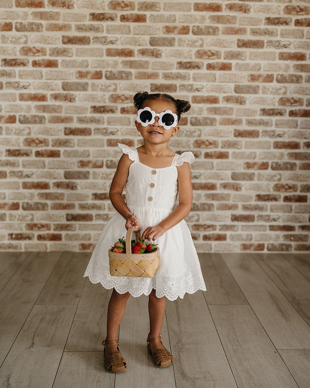 Young girl in a white dress holding a basket with flowers against a brick wall.