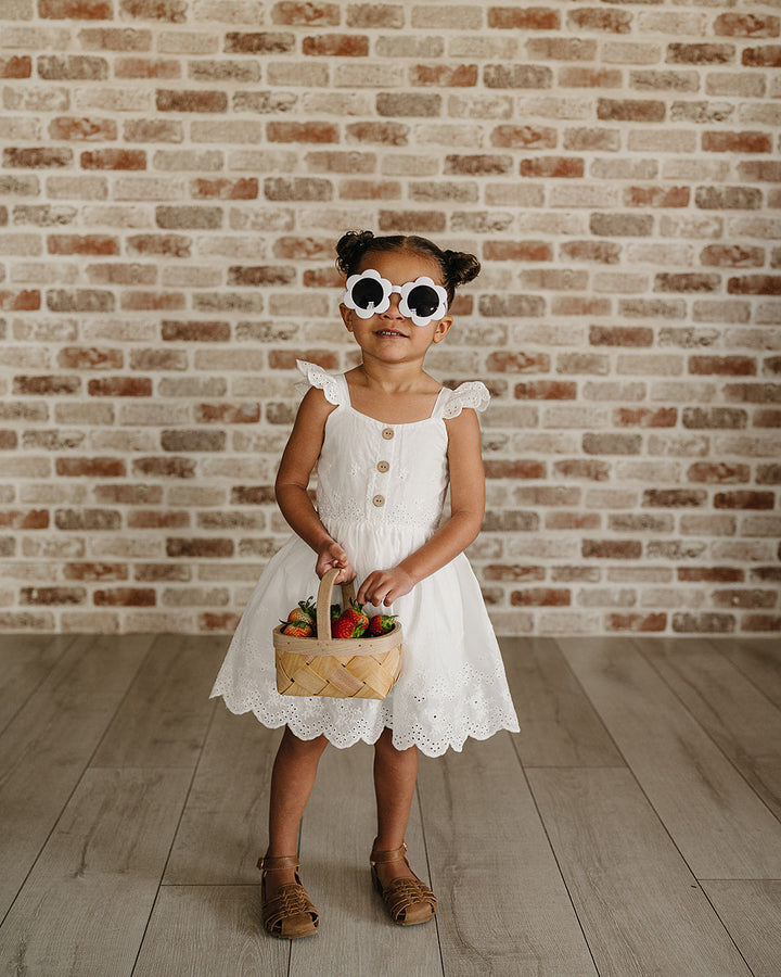 Young girl in a white dress holding a basket with flowers against a brick wall.