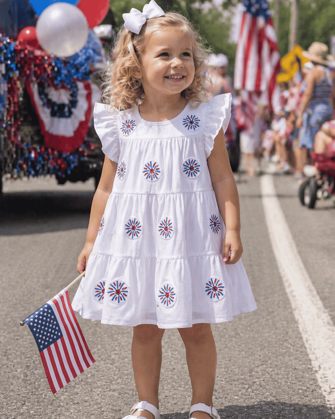 Young girl in a white dress with red, white, and blue floral patterns, holding an American flag, at a parade.