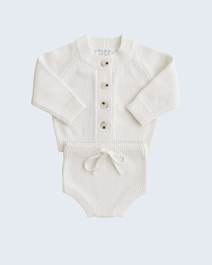 An off-white knit baby romper with long raglan sleeves, a button-up front, and a small drawstring tie at the crotch, shown on a light gray background.