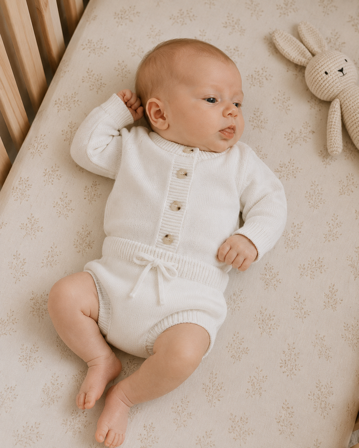 A baby lying on a patterned cream mattress, wearing a cream ribbed long-sleeve romper with button closures and a small front bow. A knitted bunny toy is visible at the top right.