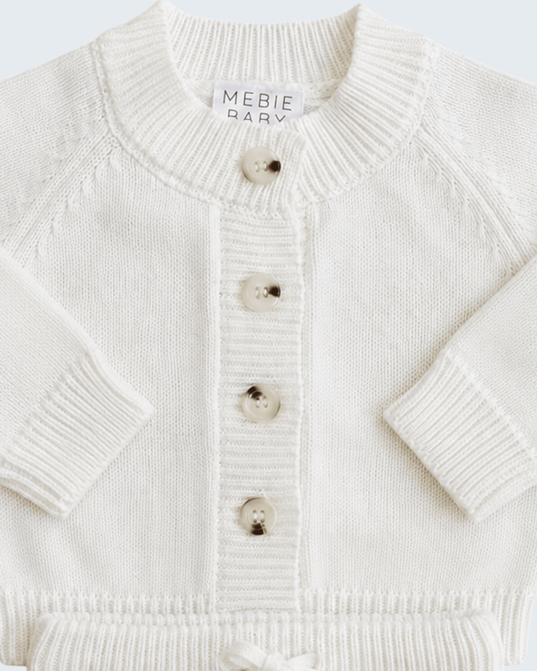 Close-up of a cream-colored knit cardigan sweater with a ribbed crew neck, button closure, and long sleeves. A brand label reading &