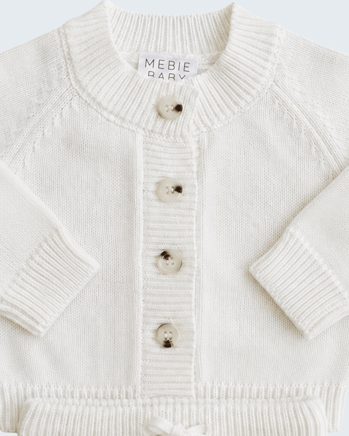 Close-up of a cream-colored knit cardigan sweater with a ribbed crew neck, button closure, and long sleeves. A brand label reading &