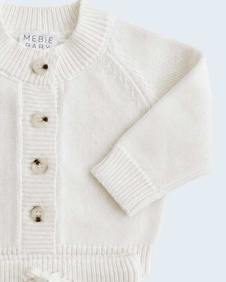 An off-white knit baby cardigan sweater with tortoiseshell buttons, shown in a close-up flat-lay view against a light gray background. The brand label &