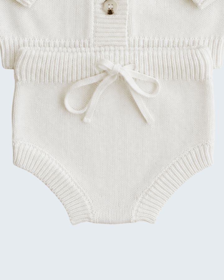 Close-up of a cream-colored ribbed knit baby bodysuit featuring a small button placket and a matching fabric bow at the waist.