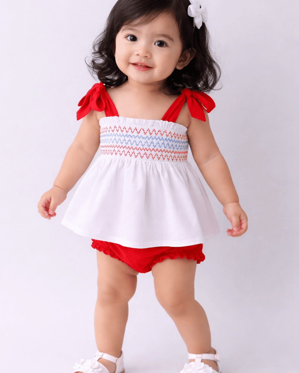 Child wearing a white and red outfit with ruffled top and bloomers on a light gray background