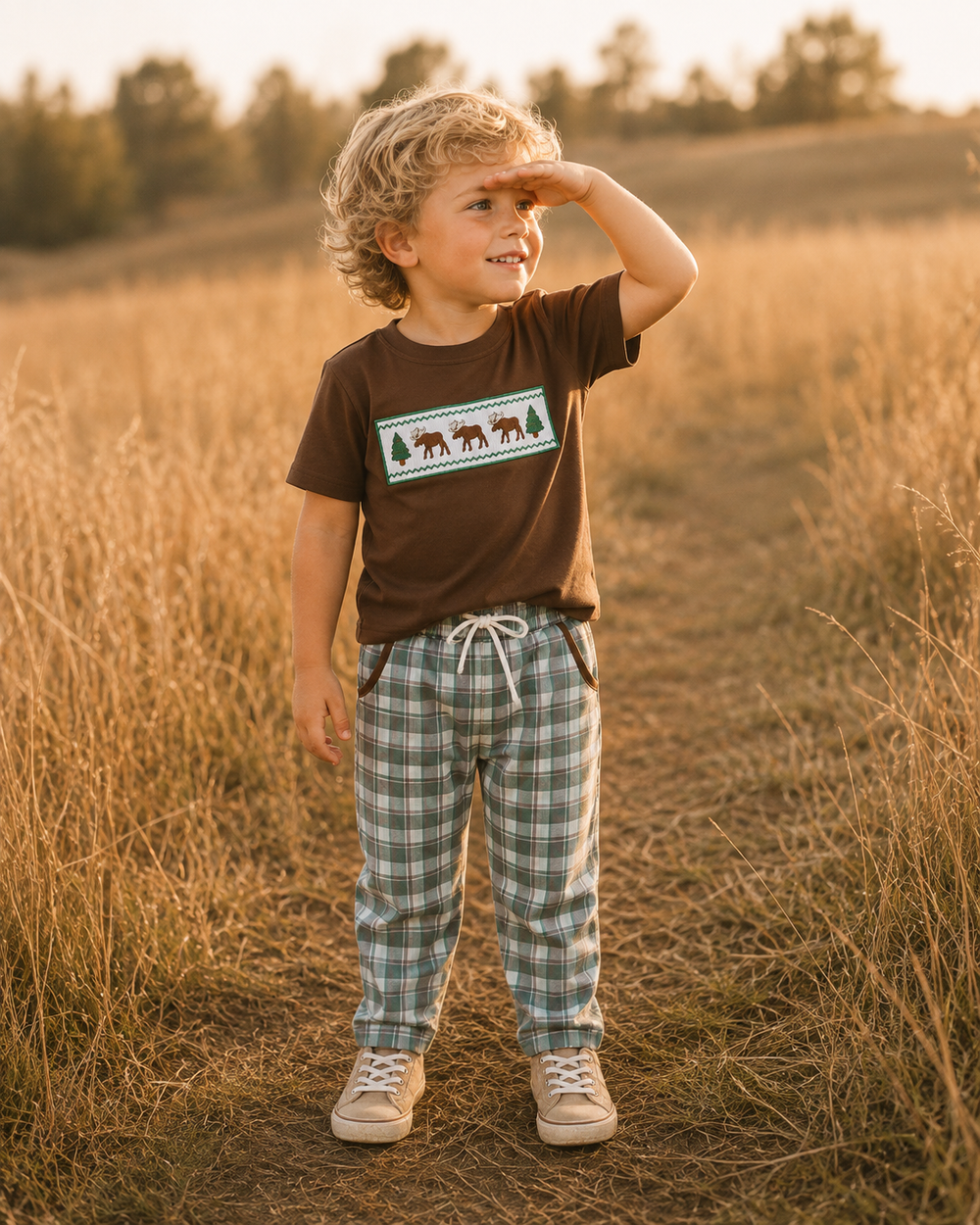 A young child standing in a dry grassy field at sunset, wearing a brown short-sleeve t-shirt with a moose graphic and green-and-white plaid pants.