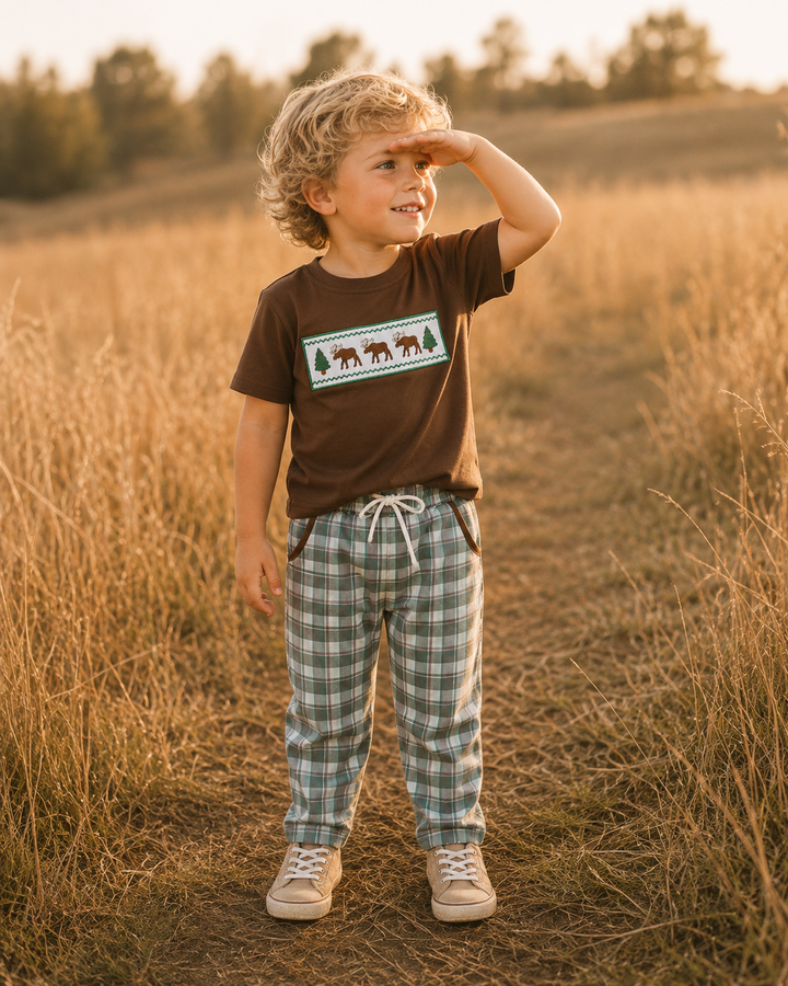 A young child standing in a dry grassy field at sunset, wearing a brown short-sleeve t-shirt with a moose graphic and green-and-white plaid pants.