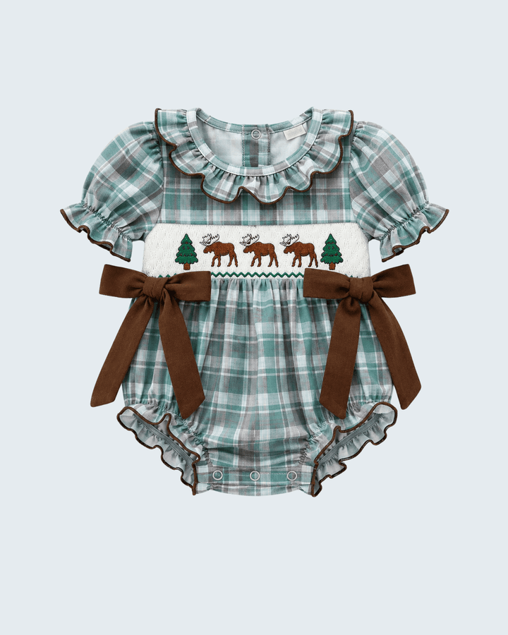 A baby romper in a green and white plaid pattern with brown trim, featuring a smocked chest panel with three brown moose, two green pine trees, and a zigzag border. It has short ruffled sleeves and two large brown ribbon bows on the sides, shown against a light gray background.