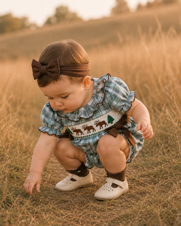 A baby girl wearing a blue and green plaid smocked romper with moose embroidery, a brown bow headband, and white shoes, crouching in a dry grass field.