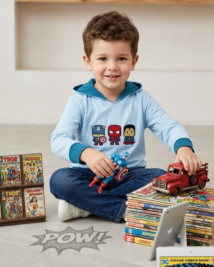 A young boy sits cross-legged on a light gray floor, wearing a light blue long-sleeve hoodie with a hood and colorful comic character prints on the front. He is surrounded by toys, comic books, and a tablet in a bright indoor room setting.