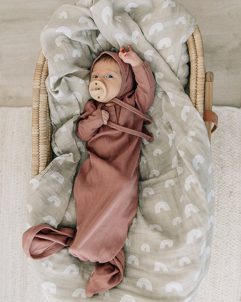 Baby wrapped in a pink knot gown with a bear toy, lying on a textured blanket in a wooden crib.