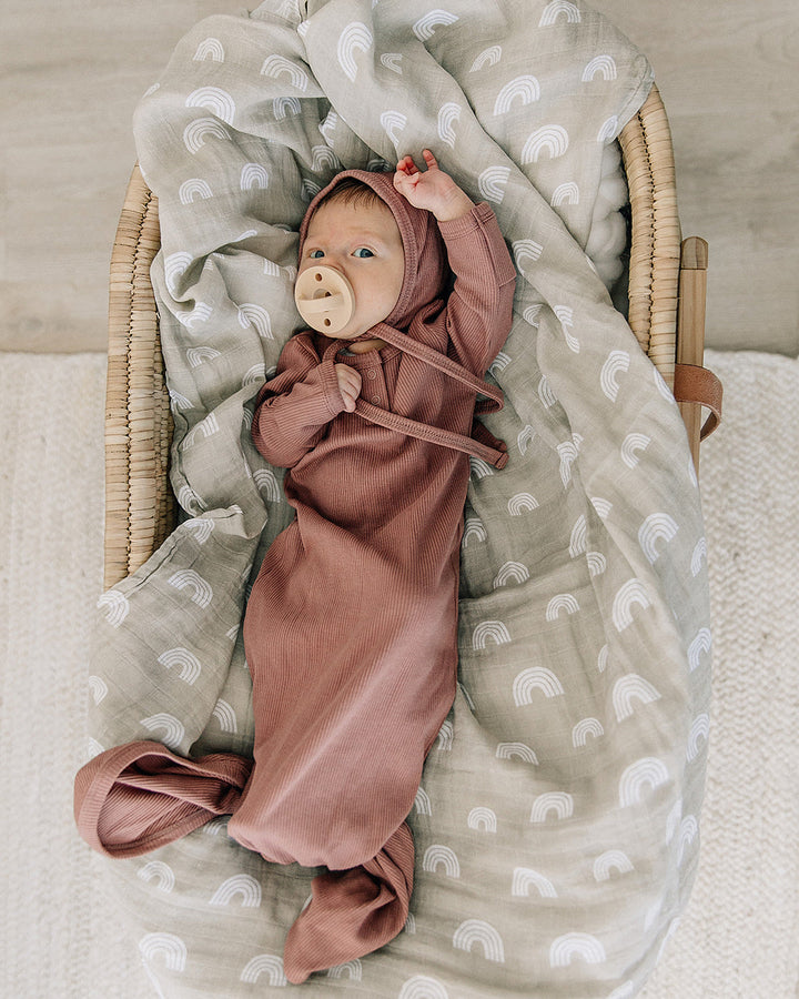 Baby wrapped in a pink knot gown with a bear toy, lying on a textured blanket in a wooden crib.