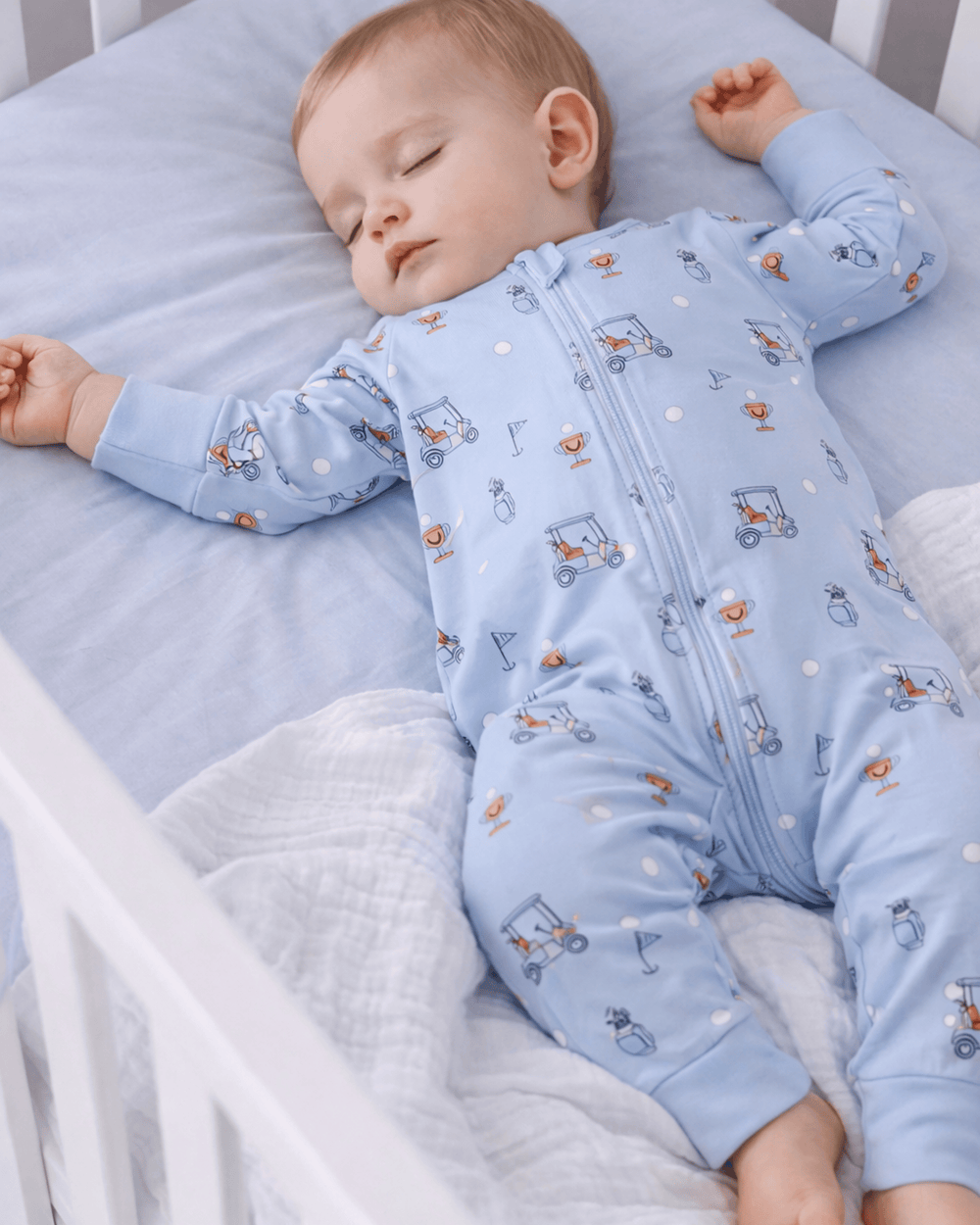 Baby sleeping in a crib wearing a blue onesie with vehicle patterns.
